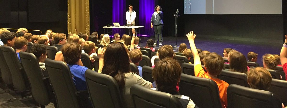 a woman standing on a stage with a microphone and a group of people in the back